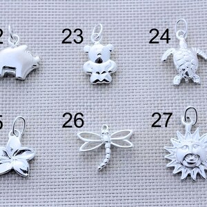 May include: A collection of six silver-toned metal charms labeled with numbers 22 through 27. The charms feature a pig, a teddy bear, a sea turtle, a five-petaled flower, a dragonfly, and a sun face with rays, each with a small jump ring attached.