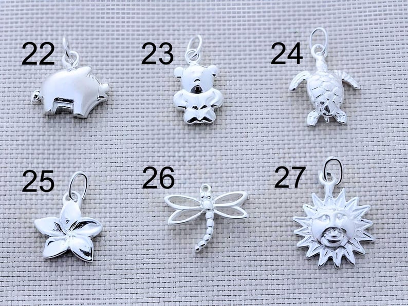 May include: A collection of six silver-toned metal charms labeled with numbers 22 through 27. The charms feature a pig, a teddy bear, a sea turtle, a five-petaled flower, a dragonfly, and a sun face with rays, each with a small jump ring attached.