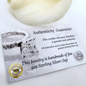 以下が含まれることがあります： 「Authenticity Guarantee」と「This certifies that your Jewellery is genuine and authentic. All material(s) used to make this product are genuine Sterling Silver Ag. This Jewelry is handmade of fine 925 Sterling Silver (Ag)」と書かれた白いカード。カードには、正方形の石がついた銀の指輪が描かれている。カードには、100%満足保証シールと本物の925スターリングシルバー保証シールも貼られている。