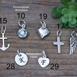 May include: A collection of seven silver-toned metal charms including an anchor, a round birthstone, a heart, a cross, a wing, and two circular and heart-shaped discs engraved with the letters K and F, each labeled with a corresponding number.