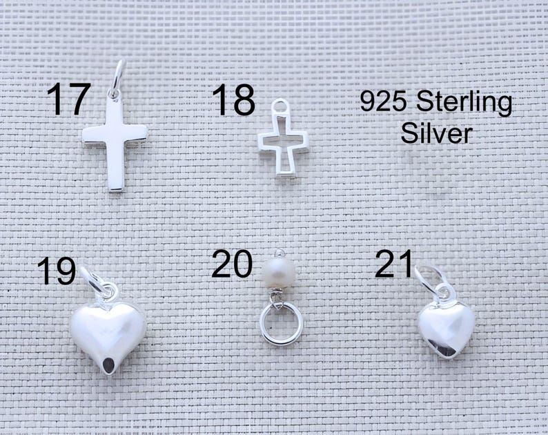 May include: A collection of 925 sterling silver jewelry charms labeled 17 through 21. The set includes a solid cross, an open-frame cross, two smooth heart charms, and a small circular charm featuring a single white pearl bead.