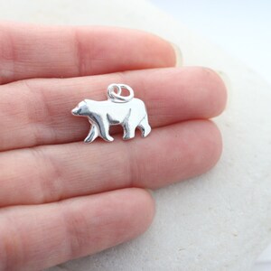 May include: A small, polished silver-toned metal charm shaped like a walking bear. The charm features a smooth, reflective surface and includes a circular jump ring attached to the top, allowing it to be easily suspended from a chain or bracelet.