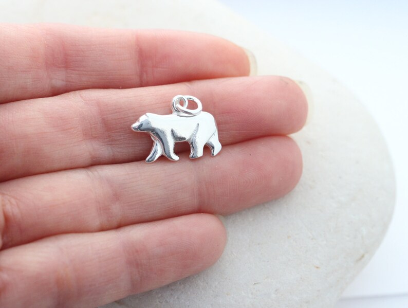 May include: A small, polished silver-toned metal charm shaped like a walking bear. The charm features a smooth, reflective surface and includes a circular jump ring attached to the top, allowing it to be easily suspended from a chain or bracelet.