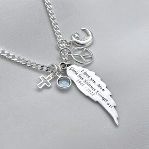 May include: Silver necklace with a charm that says "I love you, Mom Gloria Jean Holdeman Kissinger 1941 - 2022". The charm is shaped like a wing. The necklace also has a cross, a peace sign, and a crescent moon charm.