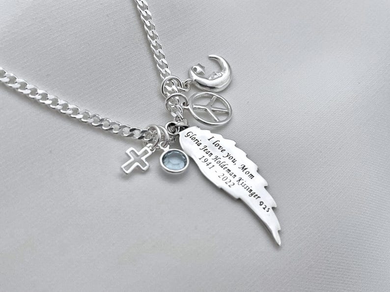 May include: Silver necklace with a charm that says "I love you, Mom Gloria Jean Holdeman Kissinger 1941 - 2022". The charm is shaped like a wing. The necklace also has a cross, a peace sign, and a crescent moon charm.