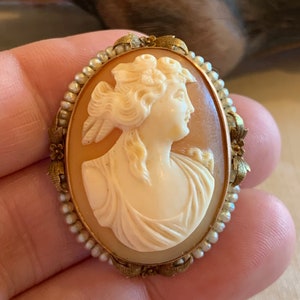 Puede incluir: Un broche de camafeo con un retrato tallado de una mujer con un tocado floral. El camafeo est&aacute; engastado en un marco dorado con un borde de perlas.