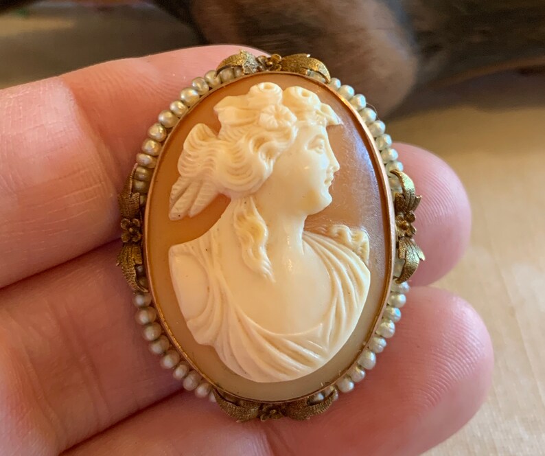 Puede incluir: Un broche de camafeo con un retrato tallado de una mujer con un tocado floral. El camafeo est&aacute; engastado en un marco dorado con un borde de perlas.