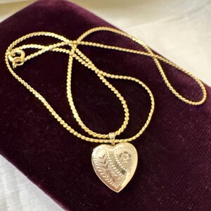 May include: A gold-colored necklace with a heart-shaped locket. The locket has an engraved floral design. The necklace chain is a twisted rope style. The locket and chain are displayed on a dark purple velvet surface.