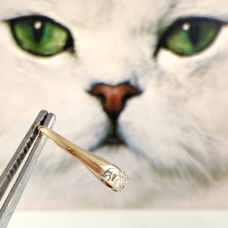 May include: A gold band ring with a detailed floral design on the top. The ring is held by a pair of tweezers.