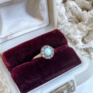 May include: A silver ring with a round opal stone surrounded by smaller clear stones. The ring is in a red velvet ring box.