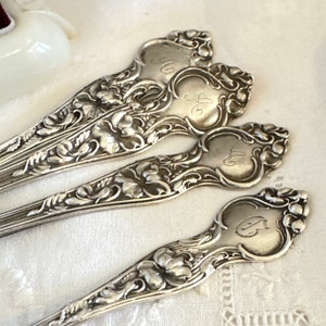 May include: Four silver spoons with ornate floral designs and engraved initials. The spoons are arranged in a row, with the handles pointing to the right.