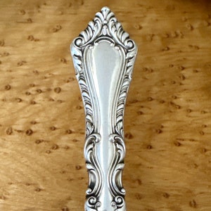 May include: Close-up of the ornate handle of a silver serving spoon. The handle is highly detailed with a decorative, floral design.
