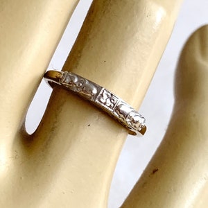 May include: A silver-toned ring with a repeating pattern of small, square designs. The ring is worn on a finger, and the details of the pattern are visible. The ring appears to be made of metal and has a polished finish.