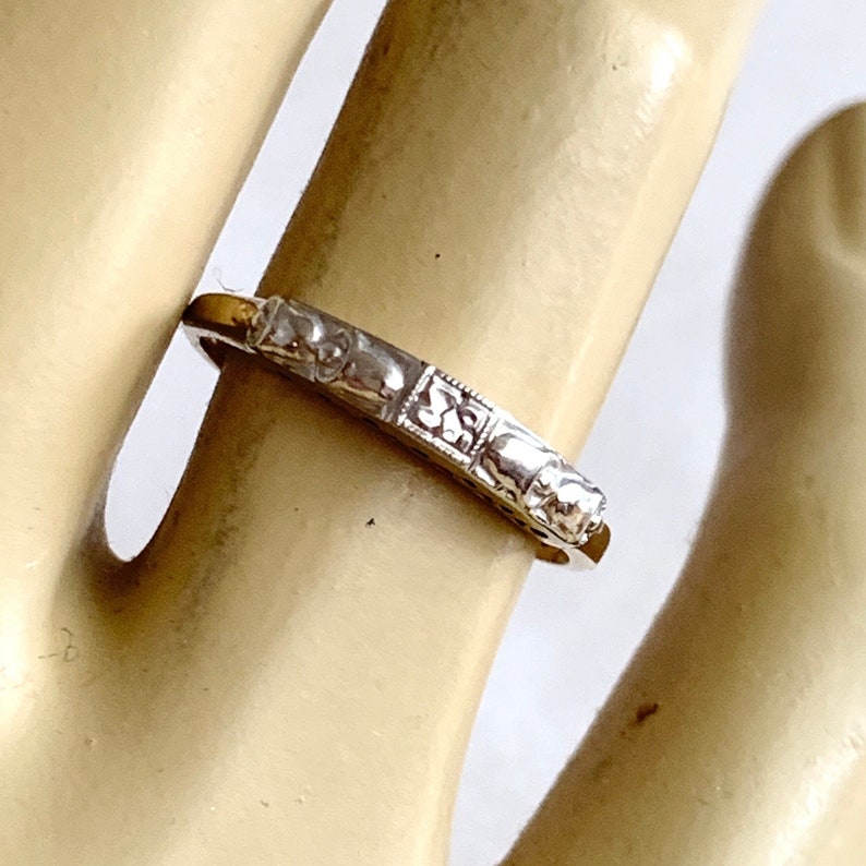 May include: A silver-toned ring with a repeating pattern of small, square designs. The ring is worn on a finger, and the details of the pattern are visible. The ring appears to be made of metal and has a polished finish.