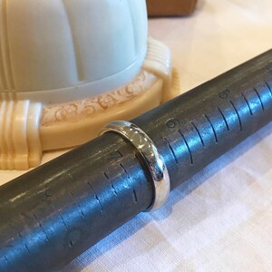 May include: A silver ring being measured on a metal ring sizer. The ring sizer has markings in inches.