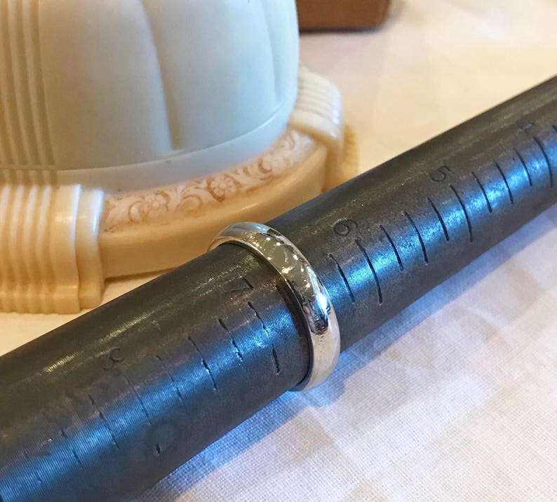 May include: A silver ring being measured on a metal ring sizer. The ring sizer has markings in inches.