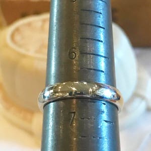 May include: A silver ring being measured on a ring sizer. The ring is a simple band style and is sized between 6 and 7.