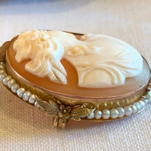 Puede incluir: Un broche de camafeo con un retrato tallado de una mujer con un vestido blanco, engastado en un marco dorado con un dise&ntilde;o de perlas y hojas.