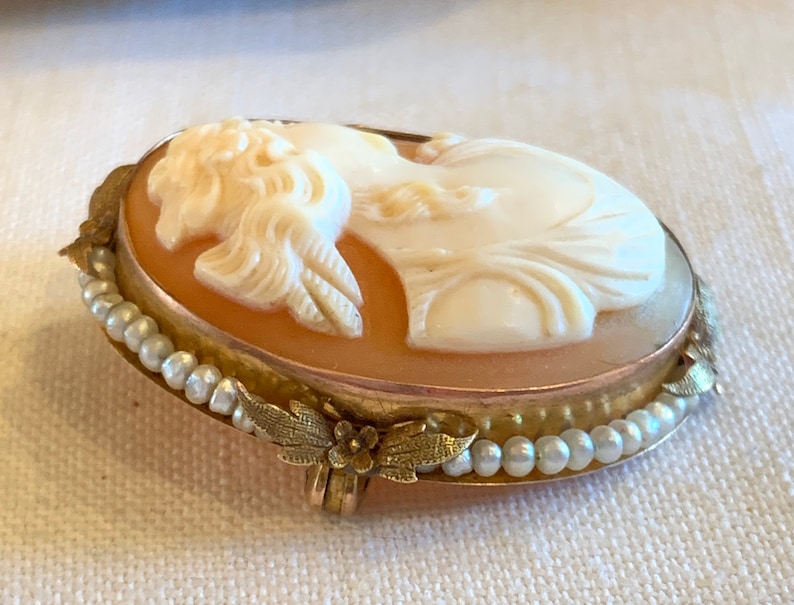 Puede incluir: Un broche de camafeo con un retrato tallado de una mujer con un vestido blanco, engastado en un marco dorado con un dise&ntilde;o de perlas y hojas.