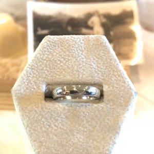 May include: A silver wedding band ring displayed on a white velvet ring box.