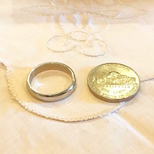 May include: A silver wedding band and a gold-colored US five-cent coin with the inscription "IN GOD WE TRUST" and "FIVE CENTS" on a white fabric background.
