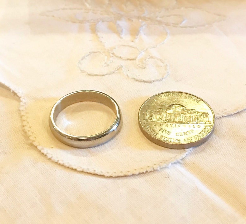 May include: A silver wedding band and a gold-colored US five-cent coin with the inscription "IN GOD WE TRUST" and "FIVE CENTS" on a white fabric background.