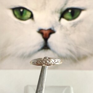 May include: A gold ring with an intricate design, featuring a central diamond-shaped stone. The ring is held by a pair of tweezers against a blurred background of a white cat's face.