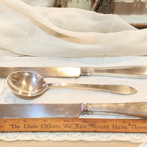 May include: Three pieces of silver flatware, including a butter knife, a soup spoon, and a dinner knife, are arranged on a white lace tablecloth. A wooden ruler with the text "Do Unto Others As You Would Have Them Do Unto You" is placed below the flatware.