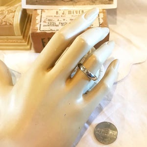 May include: A silver wedding band on a mannequin hand. The ring is simple and plain, with a smooth finish. A US quarter is shown for size comparison.