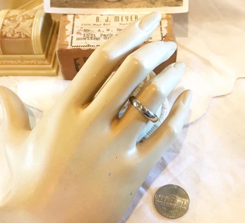 May include: A silver wedding band on a mannequin hand. The ring is simple and plain, with a smooth finish. A US quarter is shown for size comparison.