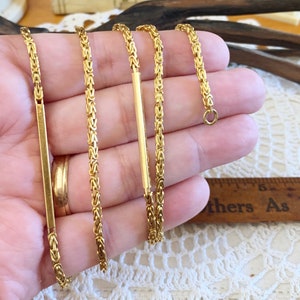 May include: A gold chain necklace with a Byzantine weave pattern. The chain is shown laid out on a hand, with a separate gold bar pendant.