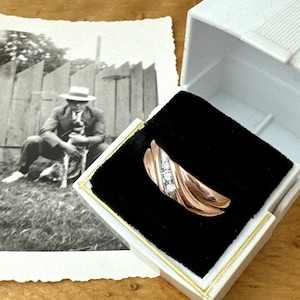 May include: A rose gold ring with three small diamonds set in a white gold band, displayed in a white jewelry box with a black velvet interior. A black and white photograph of a person with a dog is in the background.