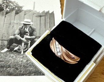 Puede incluir: Un anillo de oro rosa con tres pequeños diamantes engastados en una banda de oro blanco, exhibido en una caja de joyería blanca con un interior de terciopelo negro. Una fotografía en blanco y negro de una persona con un perro está en el fondo.