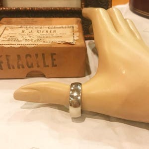 May include: A silver band ring on a mannequin hand. The ring is wide and has a smooth, polished finish. The ring is on the index finger of the hand.