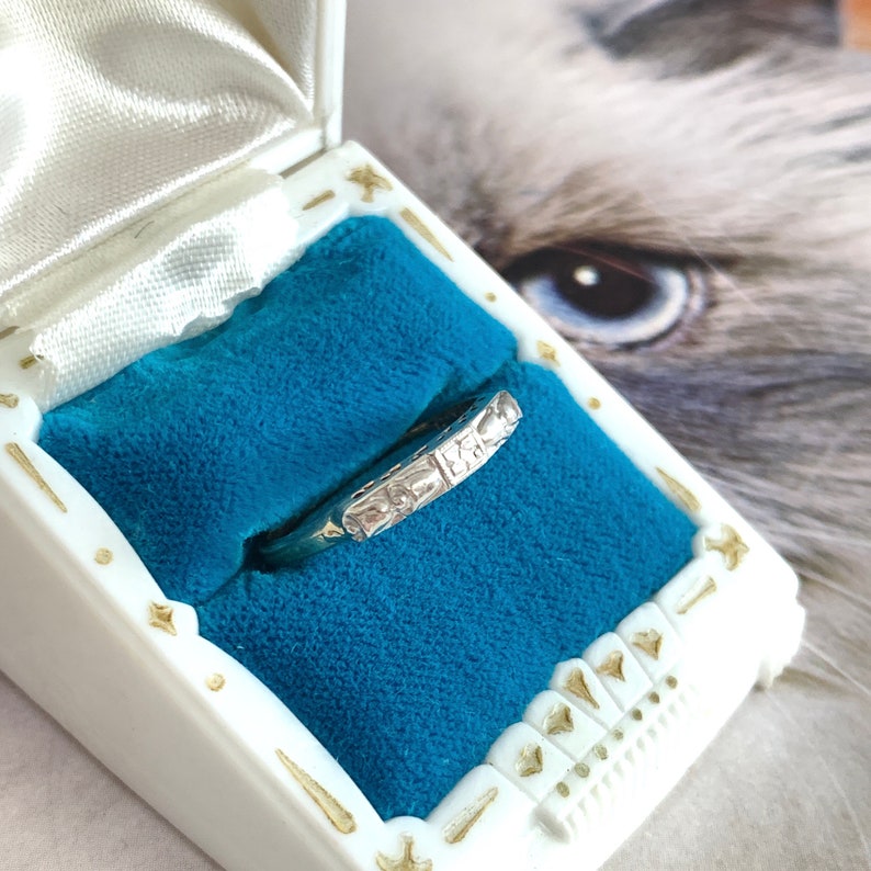 May include: A silver ring with a decorative band rests inside a white jewelry box with gold accents and a blue velvet interior. The ring's band features a repeating pattern of rectangular and square shapes. The box is open, revealing the ring.