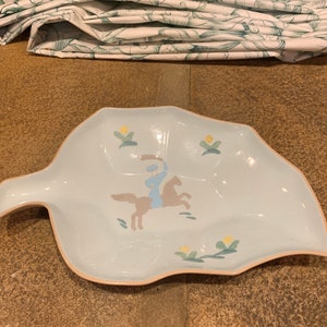 May include: A light blue ceramic leaf-shaped serving dish with a hand-painted design of a person riding a horse. The design includes yellow flowers and green leaves.