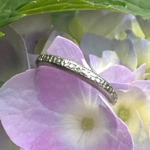 May include: A silver ring with a floral pattern rests on a purple hydrangea flower. The ring's intricate design features small, repeating flower motifs around the band. The ring is set against a backdrop of soft purple and green hues.
