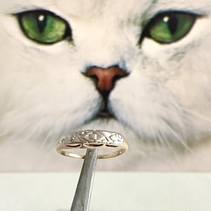 May include: A gold and white patterned band ring with a delicate design. The ring is held by a pair of tweezers.