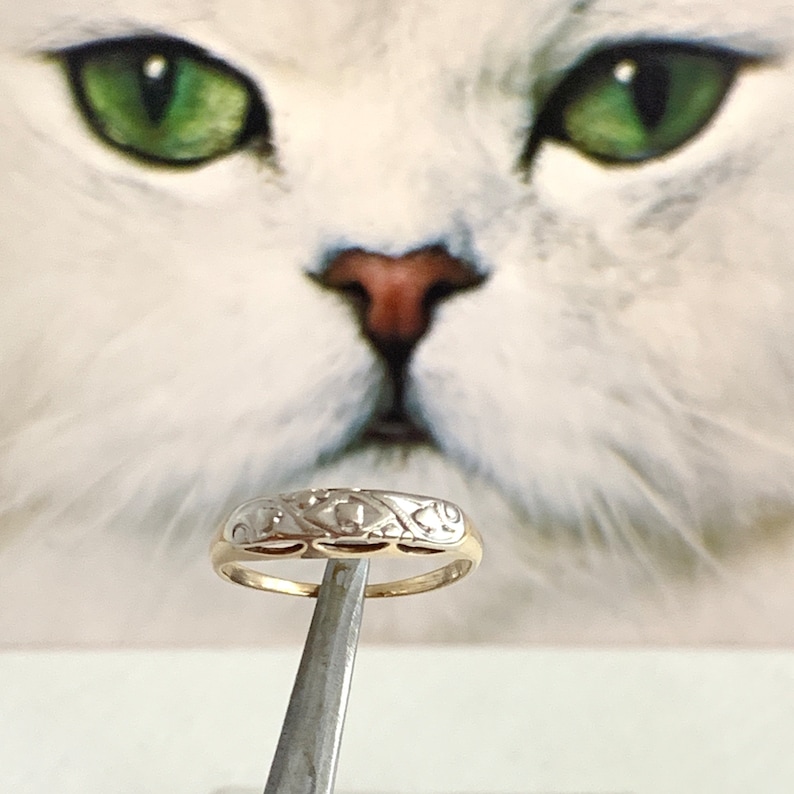 May include: A gold and white patterned band ring with a delicate design. The ring is held by a pair of tweezers.