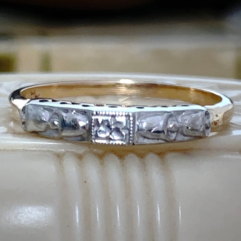 May include: A gold and silver ring with a detailed design. The ring features a band with rectangular silver sections, each with a decorative pattern. The center section has a square design. The ring is likely a piece of jewelry.