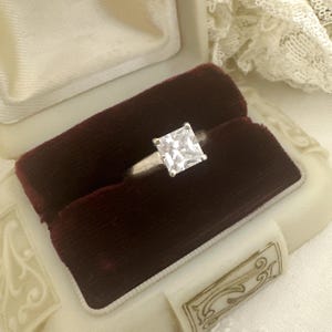 May include: A silver-toned ring featuring a single, large, square-cut clear gemstone set in a four-prong setting. The ring is displayed within a vintage-style jewelry box lined with deep burgundy velvet and cream-colored fabric.