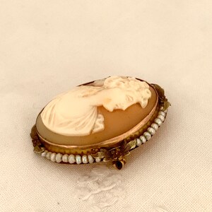 Puede incluir: Un broche de camafeo con un retrato tallado de una mujer con un borde de perlas y oro.