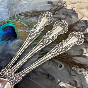 May include: Three silver spoons with ornate floral designs. The spoons are arranged in a fan shape and are resting on a bed of gray feathers.