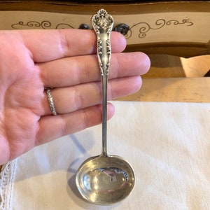 May include: A silver ladle with an ornate handle. The bowl of the ladle is round and has a smooth, polished surface.