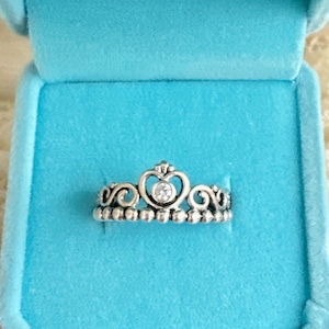 May include: A silver-toned ring in the shape of a crown, featuring a heart-shaped design with a central clear gemstone. The ring is set on a bed of small, round beads. Presented in a light blue ring box.