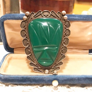 May include: A green jade mask pendant with a silver filigree frame. The mask has a carved design and is set in a silver frame with a decorative border.