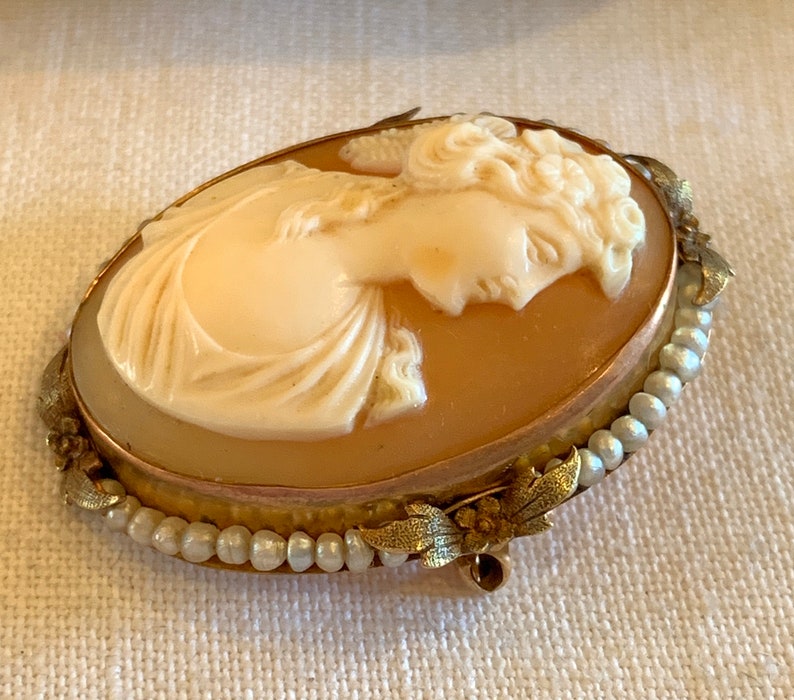 Puede incluir: Un broche de camafeo con un retrato tallado de una mujer con un vestido blanco, engastado en un marco ovalado de tono dorado con un dise&ntilde;o de perlas y flores.