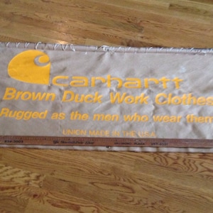 May include: A tan Carhartt banner with the text "Brown Duck Work Clothes" and "Rugged as the men who wear them." The banner also has the Carhartt logo and the text "Union Made in the USA."
