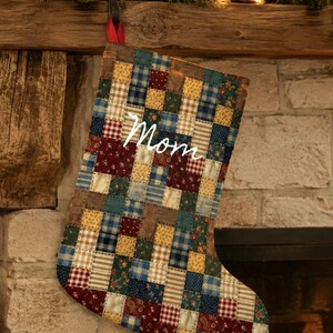 May include: A Christmas stocking made of a patchwork of small fabric squares in various patterns and colors including navy blue, burgundy, mustard yellow, and forest green. The word Mom is embroidered in white script across the center front.