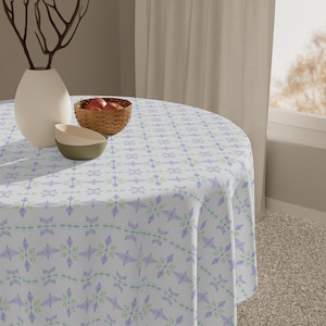 Puede incluir: Un mantel redondo con un patrón repetido de diseños florales morados y verdes sobre un fondo blanco. La mesa está puesta con un jarrón blanco, una pequeña cesta de madera con manzanas y dos cuencos.